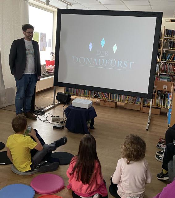 Ein Mann steht neben einer Leinwand, davor sitzen Kinder